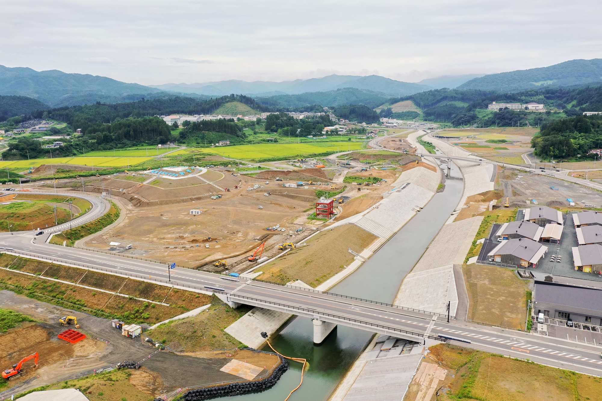 橋の上を車が通行し、下には護岸工事中の川が流れ、周囲には工事車両と整備された造成地が広がり、奥に田園と山々が見える気仙沼市内陸部の写真