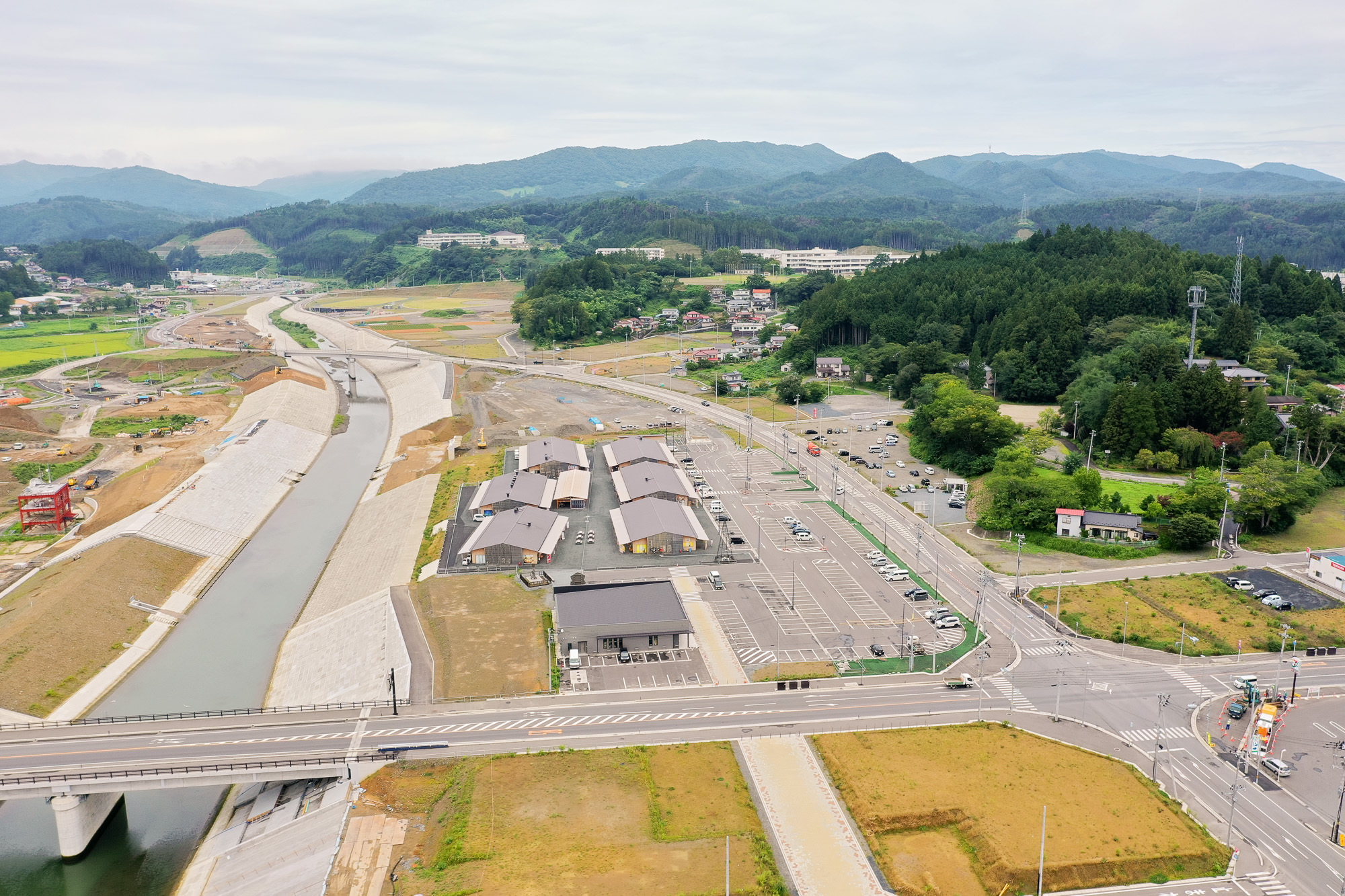 整備された護岸に囲まれた川の両側に新しい施設や駐車場が並び、奥には木々に囲まれた住宅地と山が広がる気仙沼市内陸部の写真