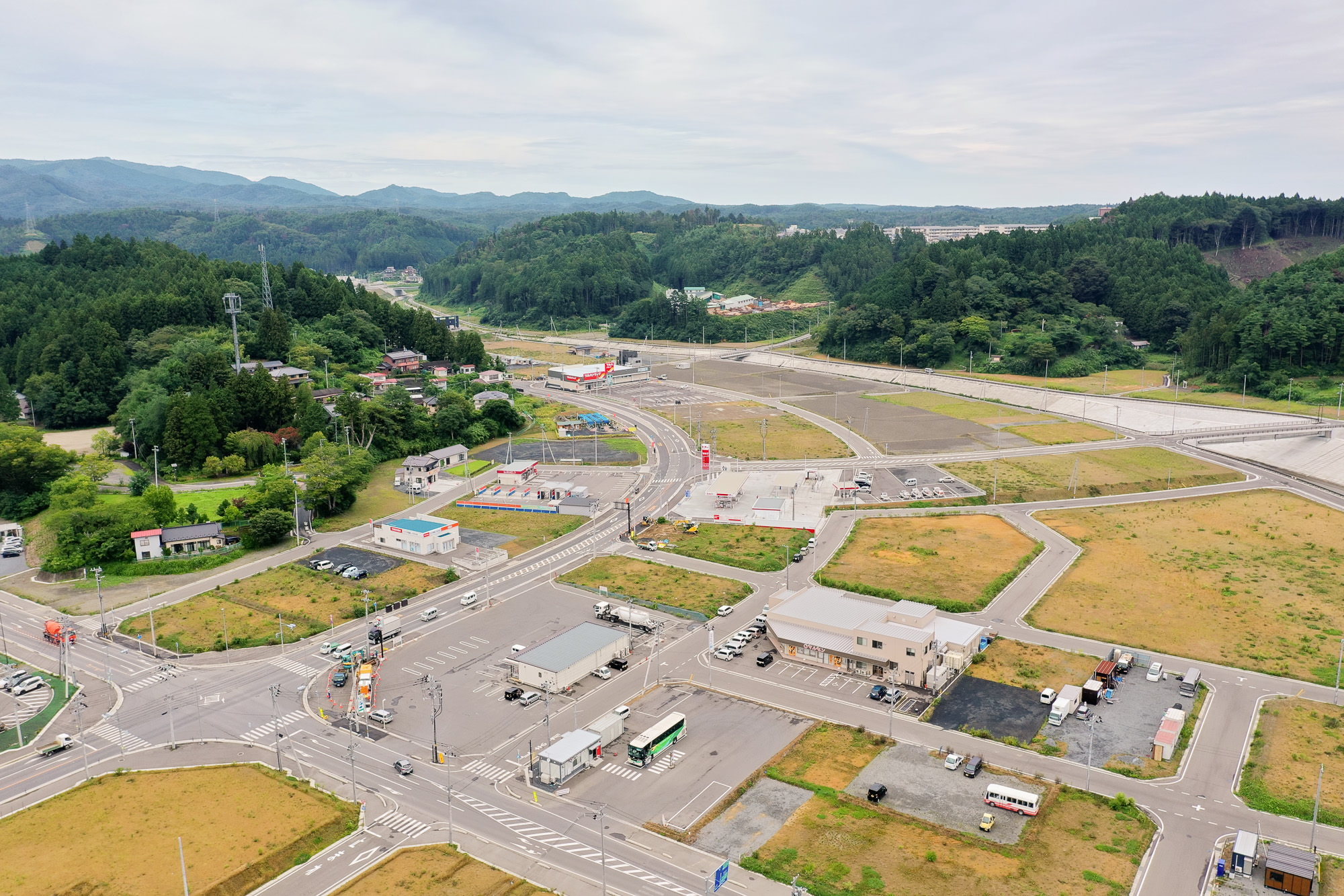 交差点周辺に商業施設や住宅が点在し、背後には木々と住宅街が広がる整備された再開発地域の市街地の写真