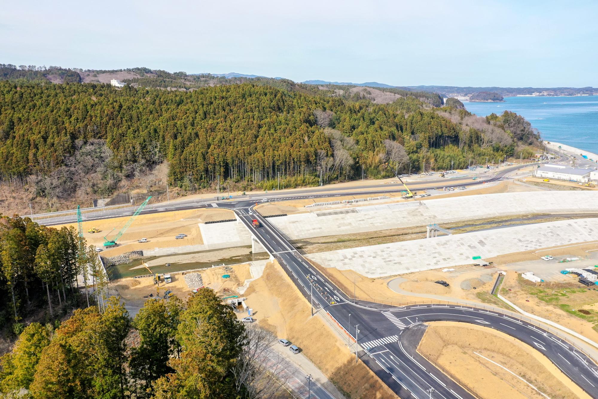 海沿いの地域にある新しく整備された道路の交差点を空から見下ろした風景で、片側には森林に覆われた丘が広がり、もう片側には海が広がっており、複数の工事車両や機材が見える開発中または完成直後のインフラ整備現場の写真