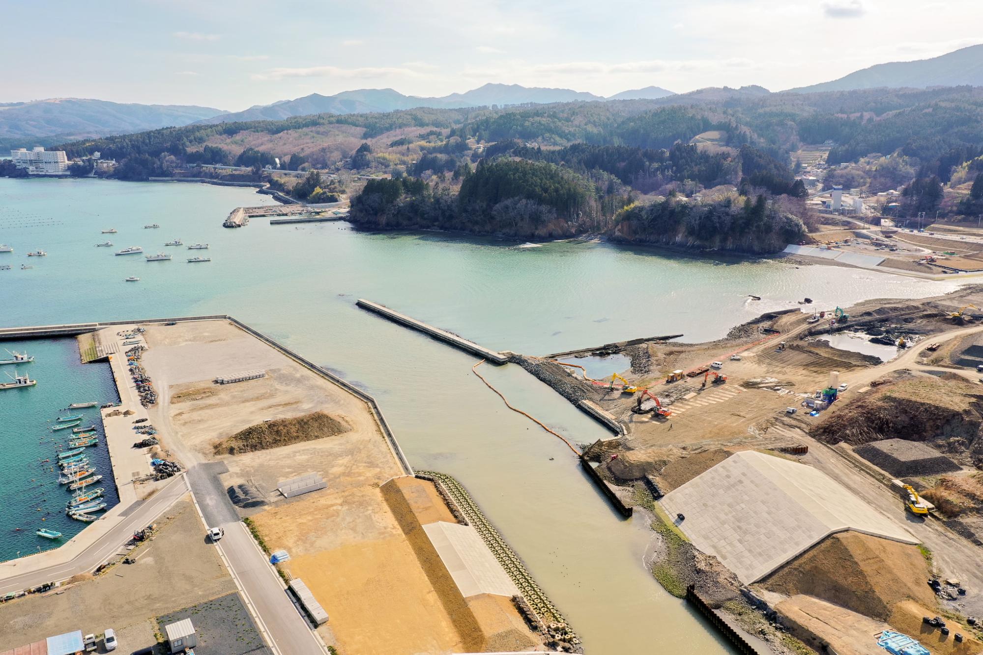 左側に複数の船が停泊する港が広がり、右側には重機と盛土が見える工事現場が展開され、背景には木々に覆われた丘陵地と点在する建物が見える海沿いの地域を空から見下ろした開発中の沿岸地域の写真