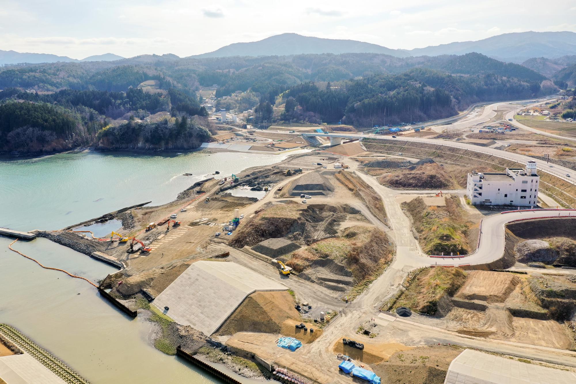 水辺の近くに広がる工事現場を空から見下ろした風景で、複数の未舗装の道路や通路、建設車両や機材が点在し、土地の造成や掘削が進められており、右側には白い建物があり、背景には丘や山々が広がる開発中の水辺地域の写真
