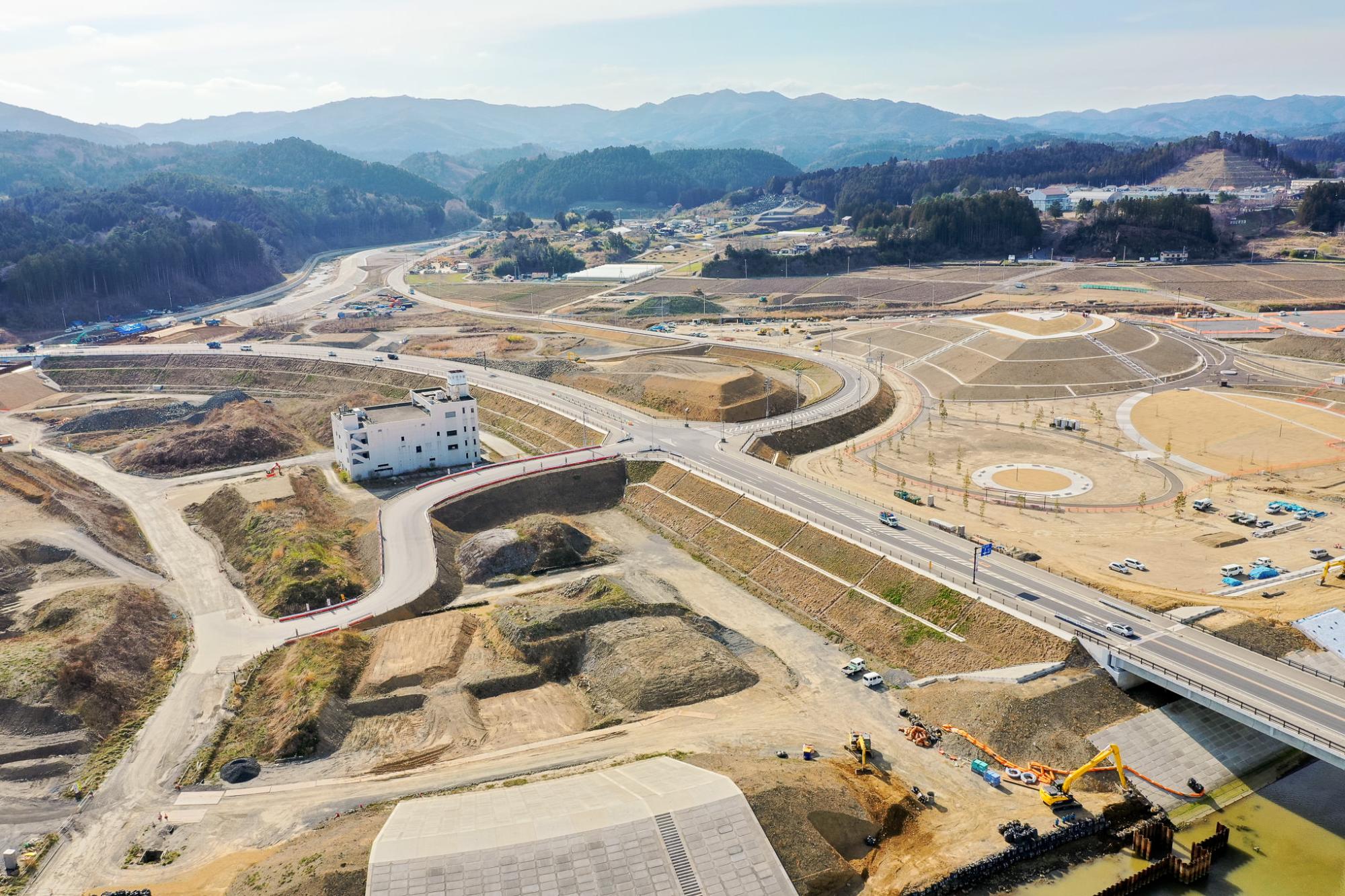 丘陵地に広がる工事現場を空から見下ろした風景で、複数の道路と橋が敷かれ、中央には白い建物があり、その周囲で建設作業や重機が稼働しており、段々状の造成地や土木工事が進行し、背景には山々と建物が立ち並ぶ地域が広がる開発中の山間地域の写真