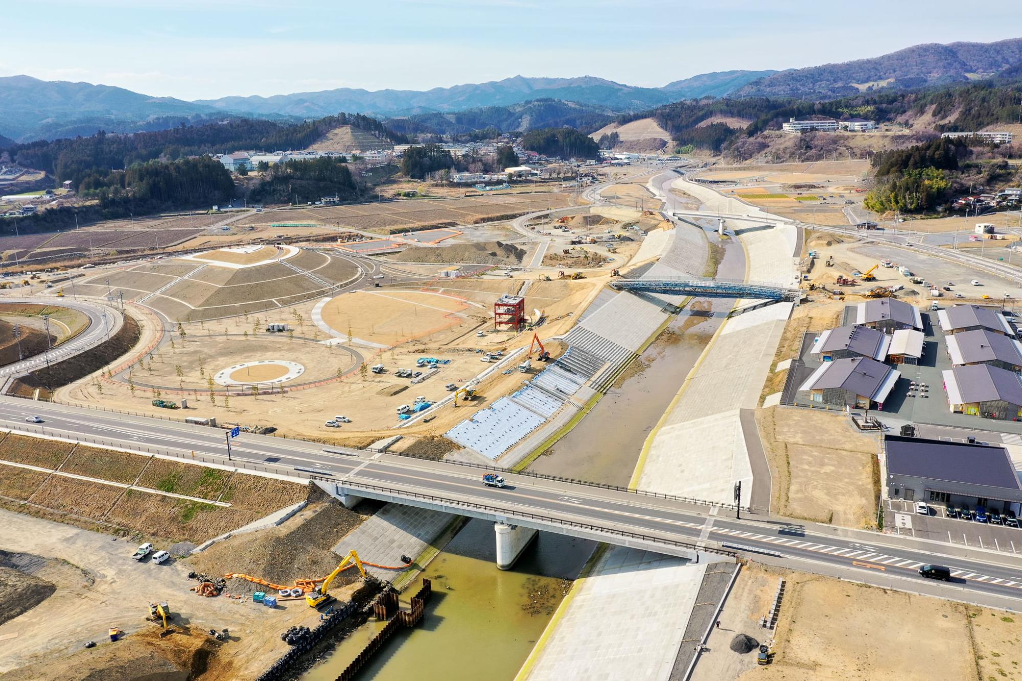 川または運河に架かる橋の周辺で複数の建設機材や車両が稼働しており、曲がりくねった道路や段々状の造成地、建設中の建物が点在し、背景には丘や山々が広がる大規模な開発または再整備が進行中の地域の写真