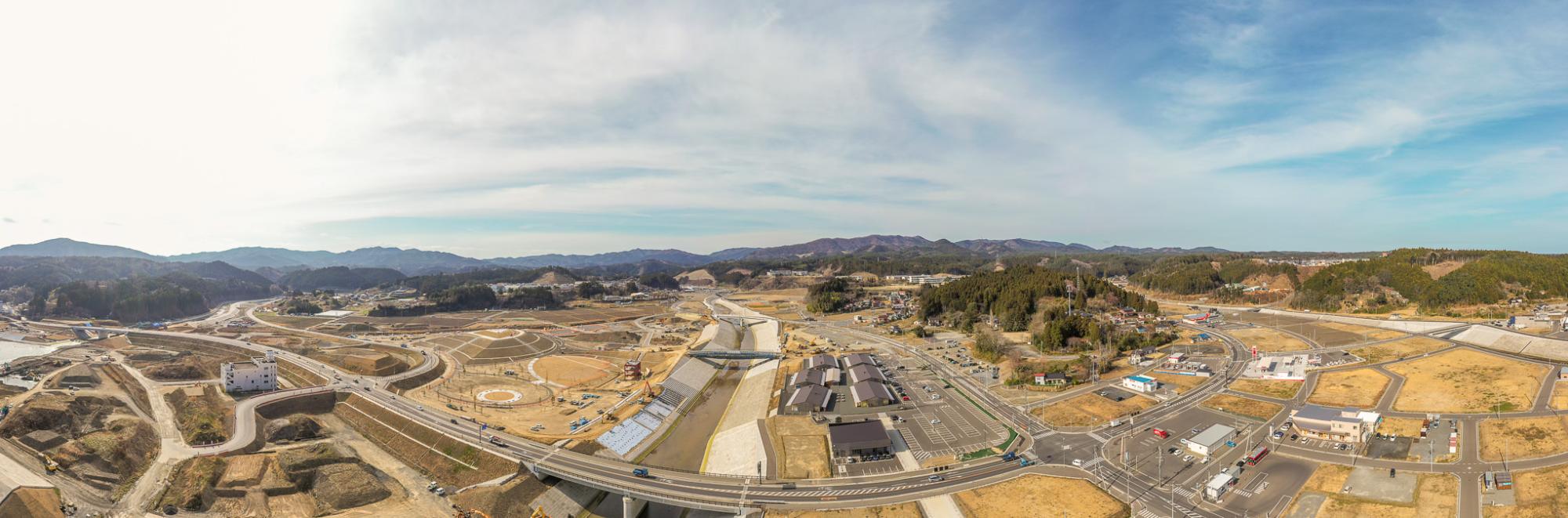 山々を背景に、自然の風景と建物・道路・橋などの人工構造物が混在する農村地域を広く見渡したパノラマ風景で、造成地やインフラ整備が進行中の様子が見られ、空は晴れて一部に雲が浮かぶ開発中の農村地域の写真