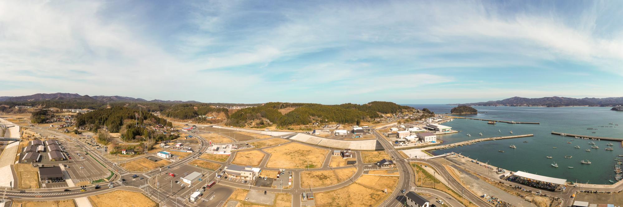 港に船が停泊し、建物や道路が広がる海沿いの町を高所から見渡したパノラマ風景で、背景には木々に覆われた丘が広がり、空には雲が点在する穏やかな沿岸都市の写真