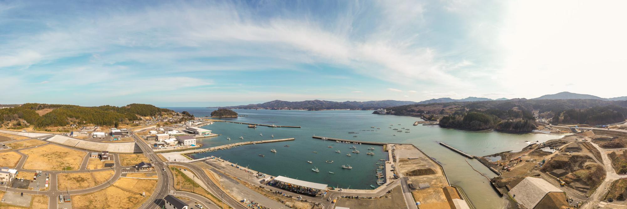 複数の小型船が停泊する港を中心に、海岸沿いに建物や構造物が並び、背景には丘や山々が広がり、空は晴れていて、手前には港周辺を結ぶ道路や通路が見える海沿いの地域を広く見渡したパノラマ風景の写真