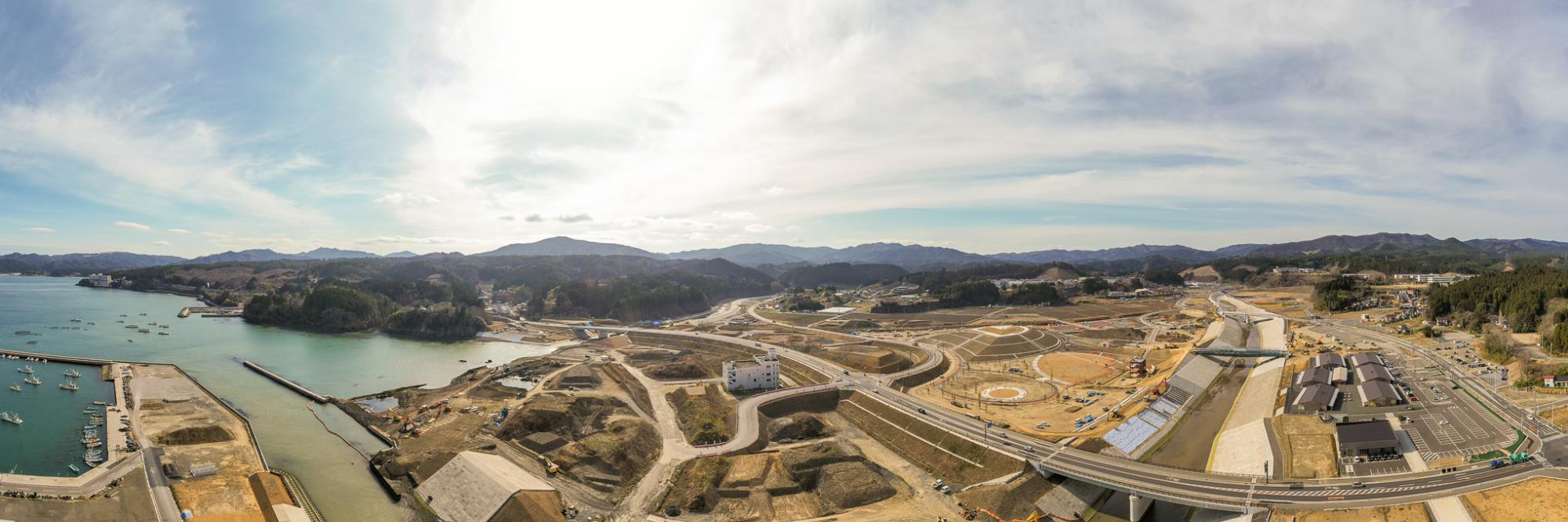 複数の道路や橋、建物が広がり、建設機材が点在する開発中の海沿いの地域を広く見渡したパノラマ風景で、港には船が停泊し、背景には丘や山々が連なり、雲が浮かぶ空から差し込む陽光が風景を照らす沿岸開発地域の写真