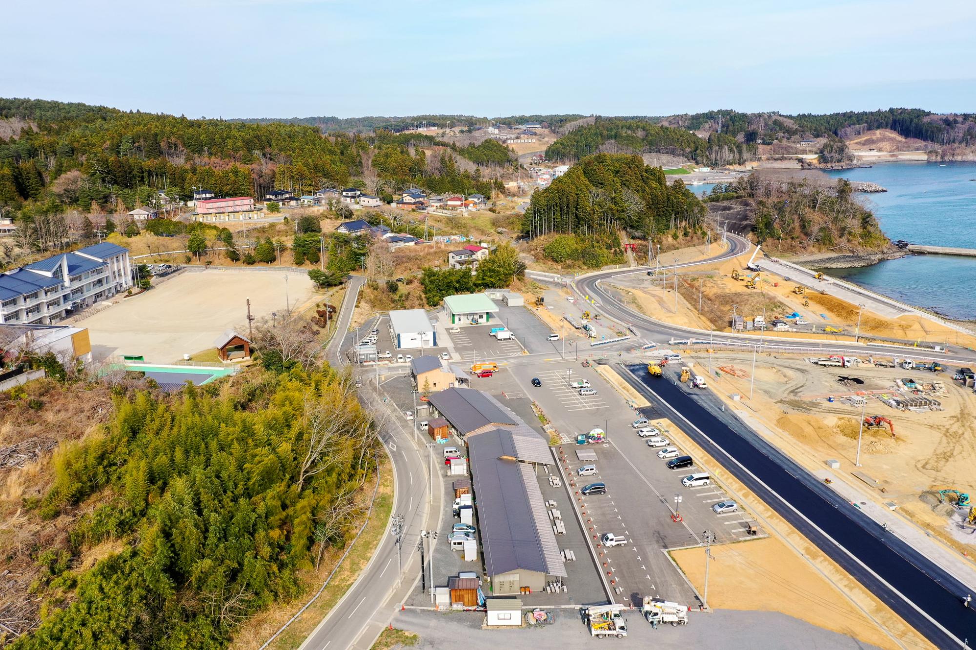 複数の建物や車両、建設機材が並ぶ広い駐車場と、新しく舗装された道路が海岸線に沿って走り、右側に海が広がり、背景には森林に覆われた丘と点在する住宅が見える自然と都市機能が融合した沿岸地域の写真