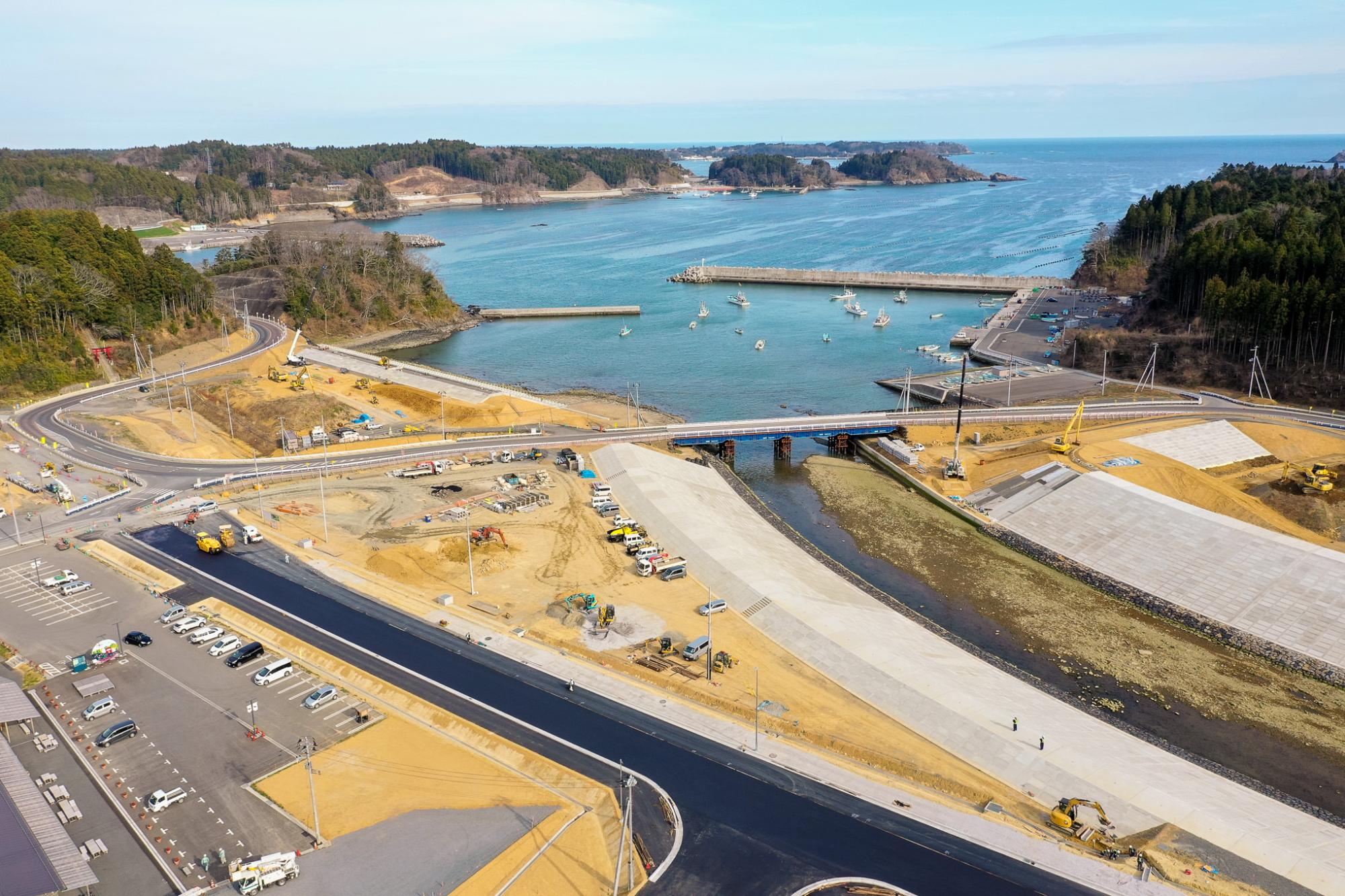 港に複数の船が浮かび、水路には橋が架かり、陸地には建設車両や機材が点在する工事現場が広がり、周囲には森林に覆われた丘と開けた土地があり、インフラ整備が進む海沿いの地域の写真