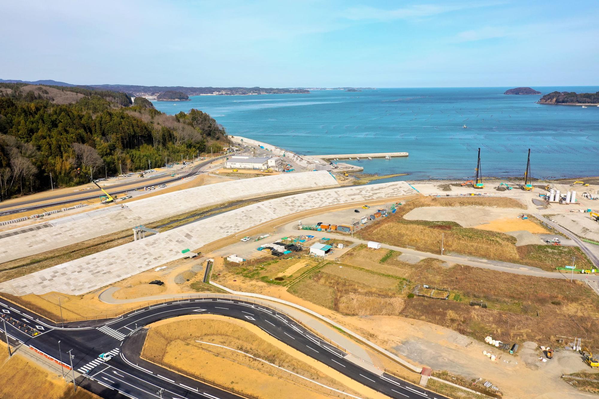 海沿いの広大な工事現場を高所から見下ろした風景で、複数の造成地と建設資材・機材が点在し、海には防波堤が突き出し、背景には木々に覆われた丘が広がり、手前には舗装された新しい道路が曲線を描いて走っている穏やかな海と遠くの島々が見える沿岸開発地域の写真