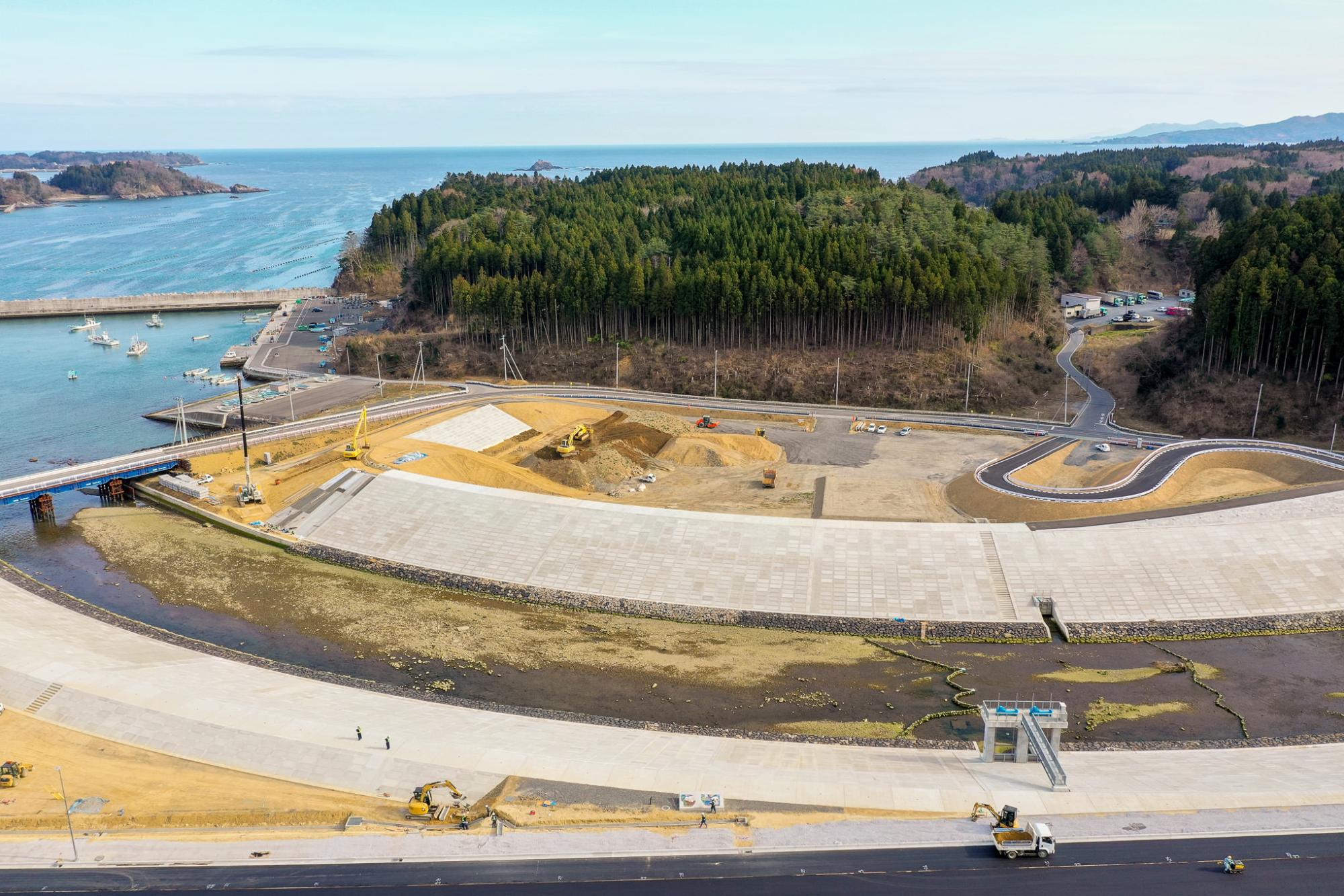 広い舗装地に建設車両や機材が点在し、背景には森林に覆われた丘と海が広がり、海には数隻の船が停泊する小さな港が見え、晴天のもとで工事が進む海沿いの地域の写真