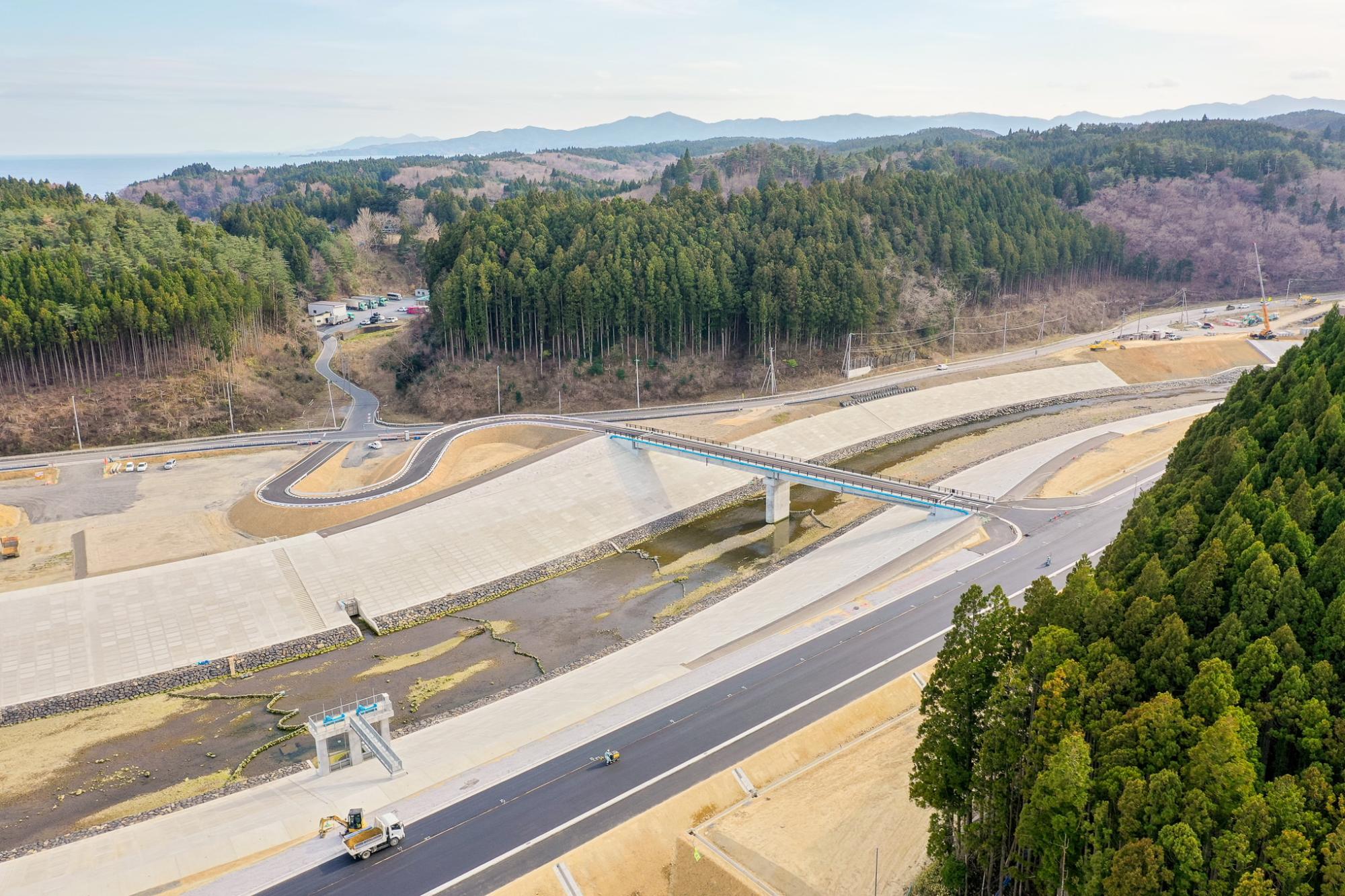 川または運河に沿ってコンクリートの護岸が整備され、その横を新しく建設された道路が走り、橋が川をまたいで両側の道路をつなぎ、周囲には密集した森林と起伏のある地形が広がり、背景には山々が見える晴天の農村地域の写真