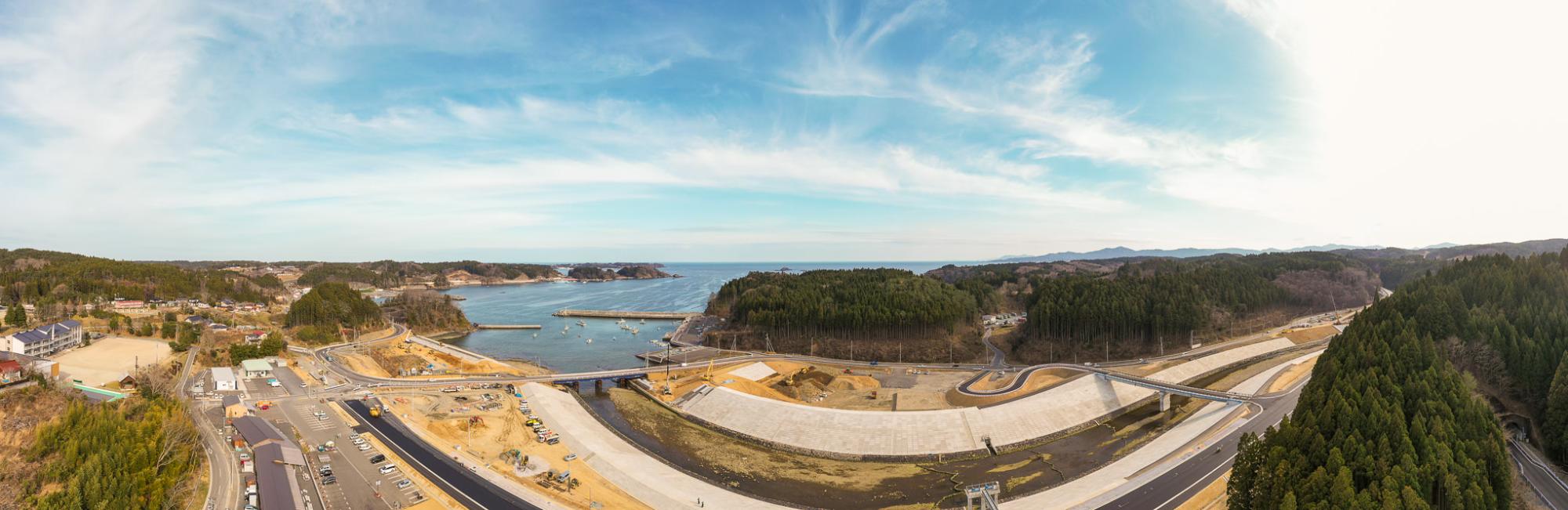 道路や駐車場、工事現場が広がる海沿いの地域を広く見渡したパノラマ風景で、背景には森林や丘陵、入り江を含む海が広がり、橋や各種インフラが点在する自然と人工構造物が融合した沿岸地域の写真
