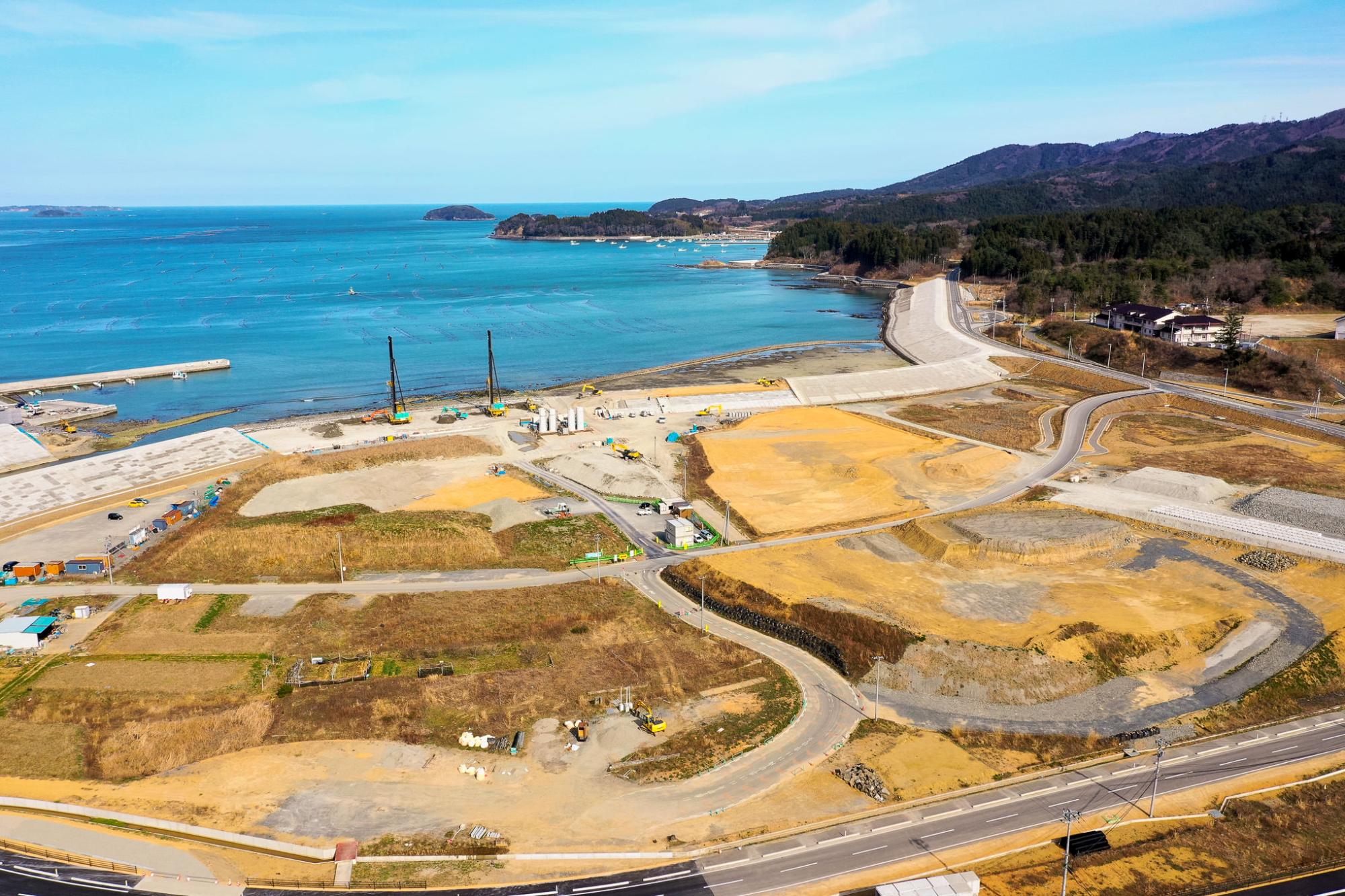 海沿いの地域で複数の造成地と道路が整備され、海岸には防波堤が築かれ、クレーンや建設機材が水辺近くに配置されており、背景には海と小さな島々、森林に覆われた丘が広がる沿岸開発中の地域の写真
