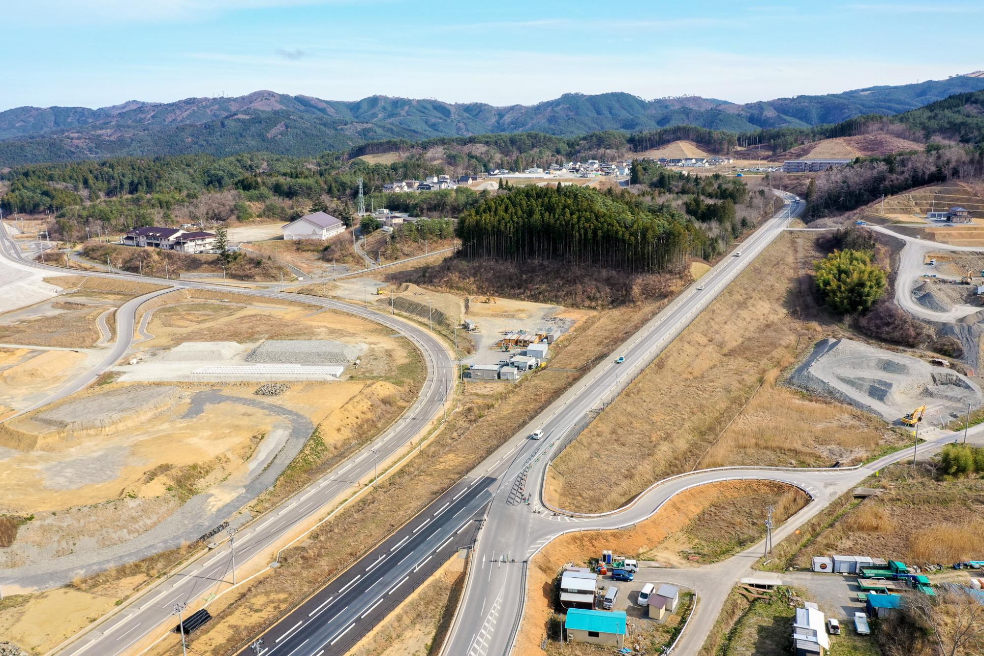 山々を背景に、曲がりくねった道路といくつかの建物、森林が広がる農村地域を空から見下ろした風景で、複数車線の幹線道路には車両が走行しており、所々に造成地や工事現場が見える開発が進む地方の写真