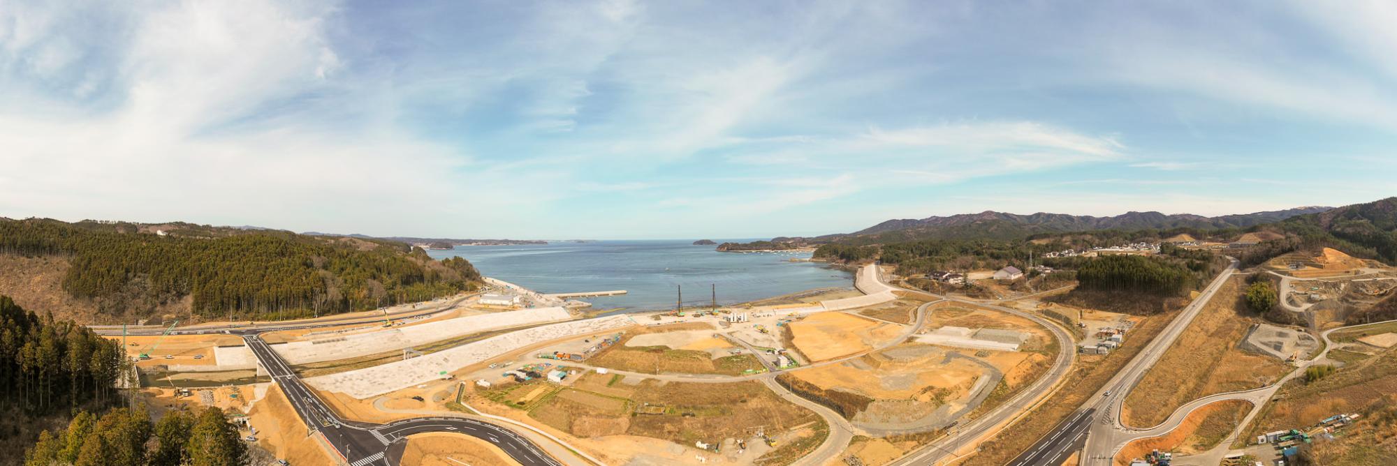 道路や工事現場、緑地が点在する沿岸地域を高所から見下ろしたパノラマ風景で、背景には海が広がり、空には少し雲が浮かぶ晴れた日の海岸地域の写真