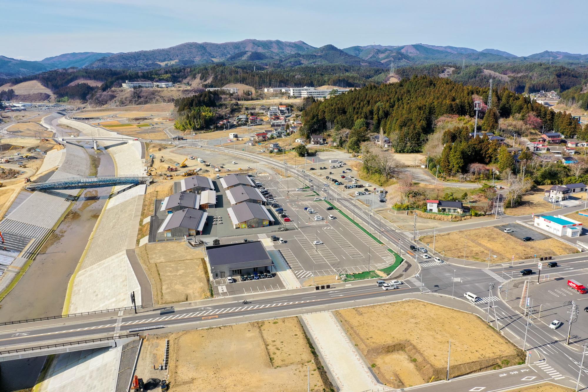 複数の建物や駐車場、道路、堤防に囲まれた川が広がる農村地域を空から見下ろした風景で、背景には丘や森林が広がり、橋や交差点などのインフラが整備された自然と人工構造物が調和する地域の写真