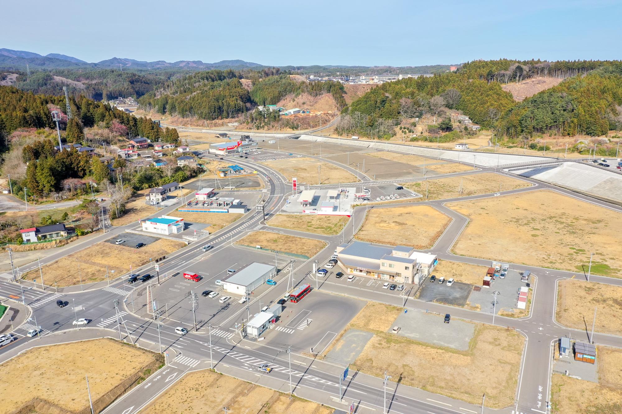 複数の道路と交差点、建物、広い空き地が広がる農村地域を空から見下ろした風景で、道路には車両が走行しており、背景には丘や木々が広がる交通インフラと自然が共存する地域の写真
