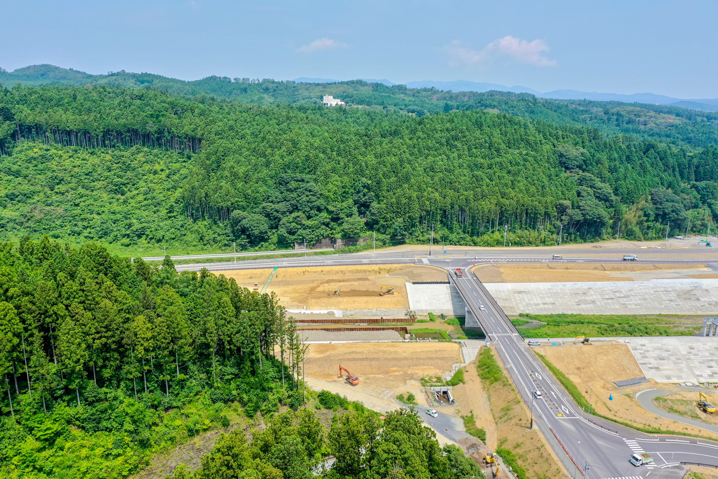 手前に複数の道路が交差する工事現場が広がり、重機が点在する整地された土地が見え、背景には森林が広がり、空には少し雲が浮かぶ晴れた日の風景の写真