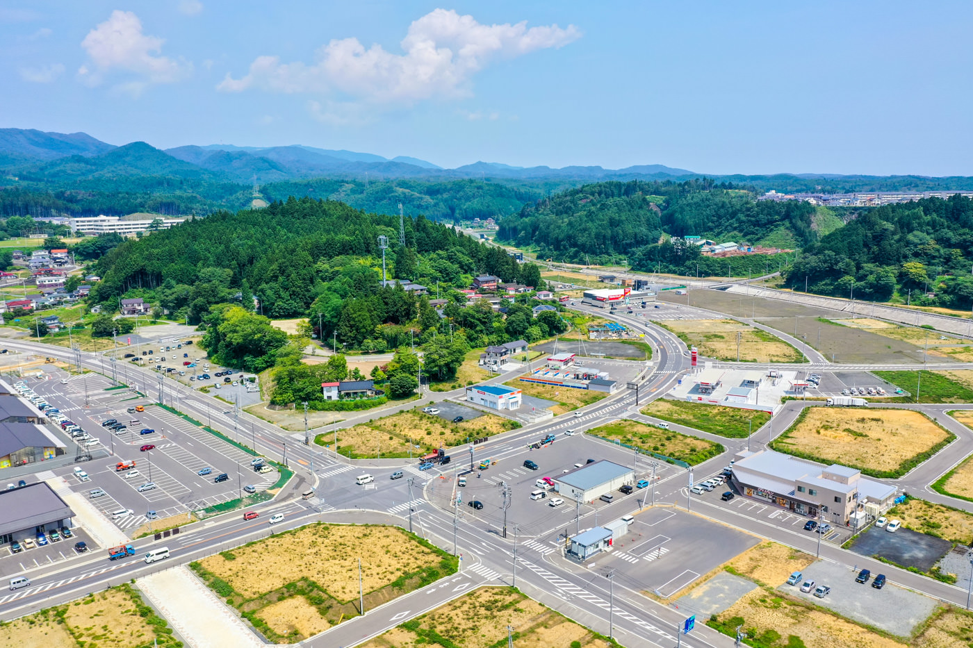 複数の道路が交差し、建物や駐車場、緑地が点在する郊外地域を上空から見下ろした風景で、車が道路や駐車場に見られ、背景には森林に覆われた丘や山々が広がる晴天の写真