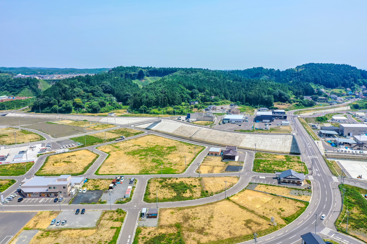 複数の区画や建物、道路が広がる半農村地域を上空から見下ろした風景で、緑地や空き地が点在し、遠くには森林に覆われた丘が広がる晴天の写真