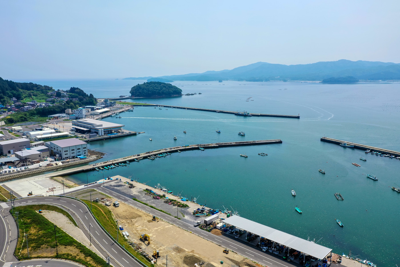 港に複数の桟橋と船が並び、海岸沿いには建物が立ち並び、手前には複数車線の道路が走り、背景には海を挟んで島と山々が広がる晴天の海辺の風景の写真