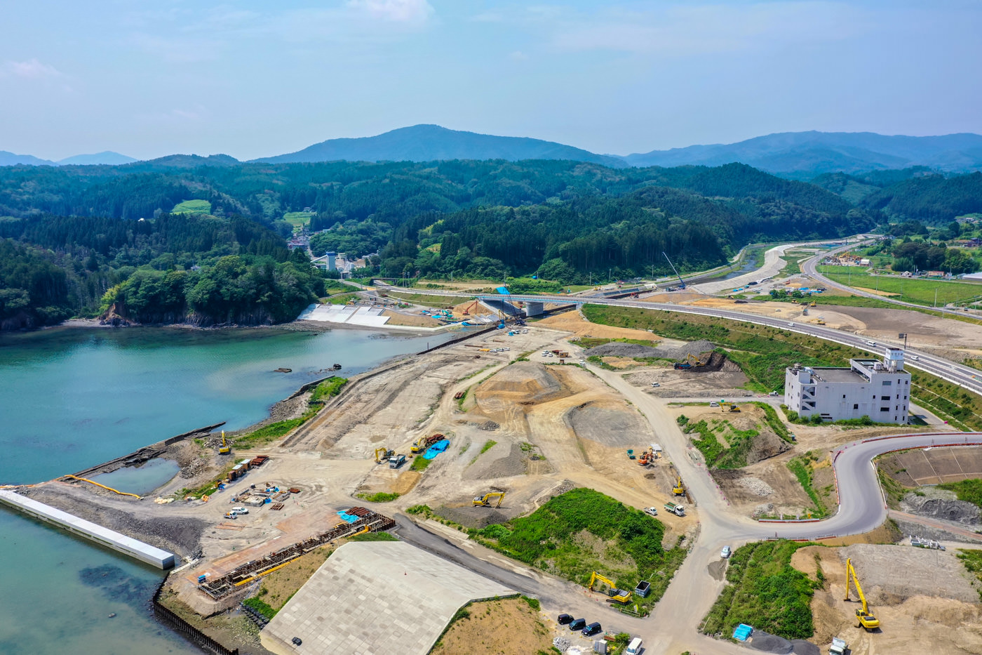 緑の山々に囲まれた湖のほとりで、複数の建設車両と機材が並ぶ広い工事現場が広がっており、青空の下に道路や建物も見える風景の写真