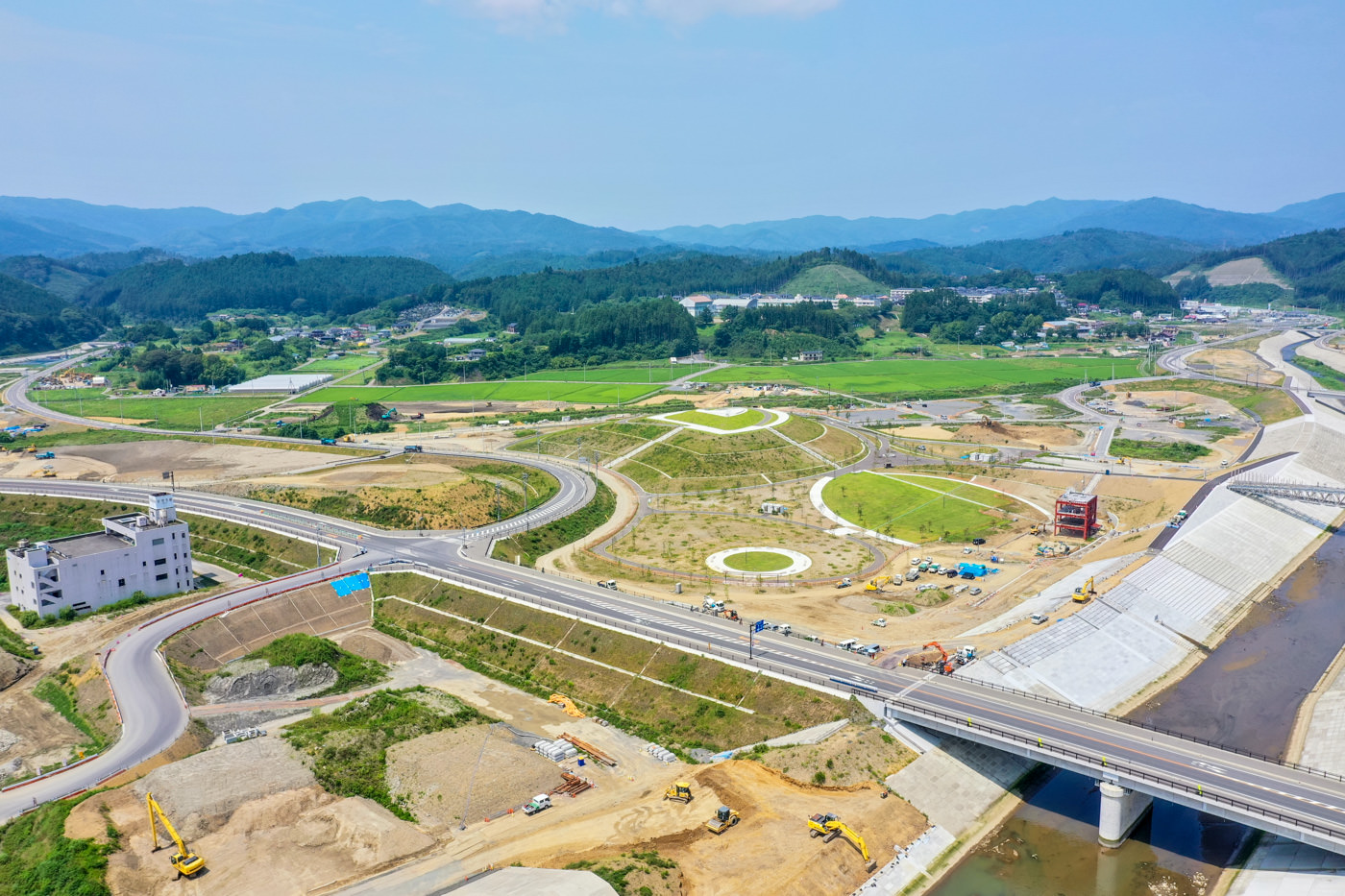 川にかかる橋と複数の道路や通路が整備中の工事現場を上空から見下ろした風景で、周囲には緑地や畑が広がり、背景には山や丘がそびえ、現場には建設機械が点在している写真