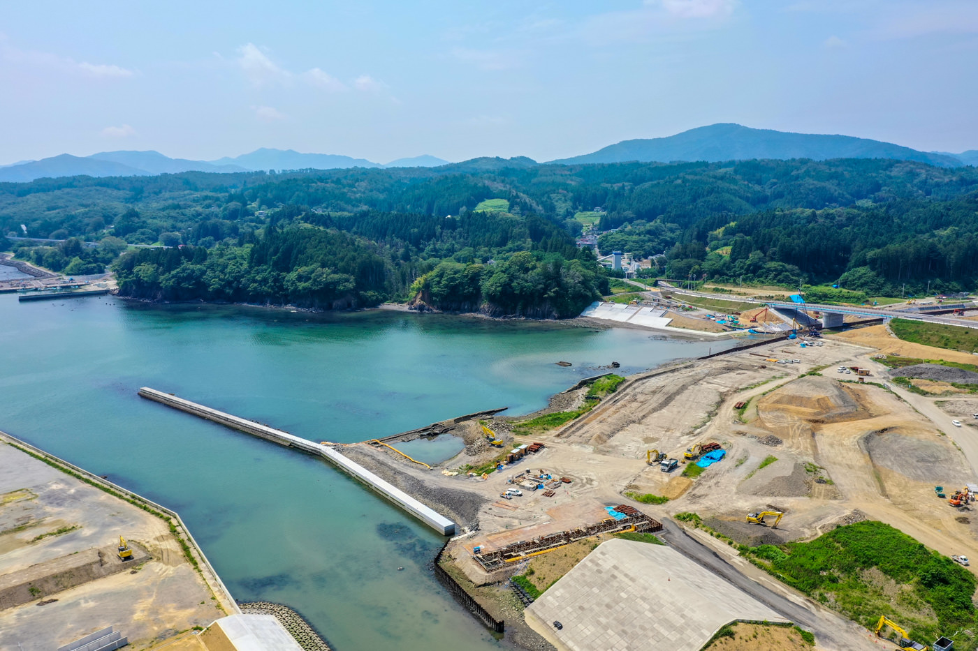 山々に囲まれた湾の一部で、防波堤が湾内に設置されており、その周囲には工事現場や建設中の構造物が見える風景の写真