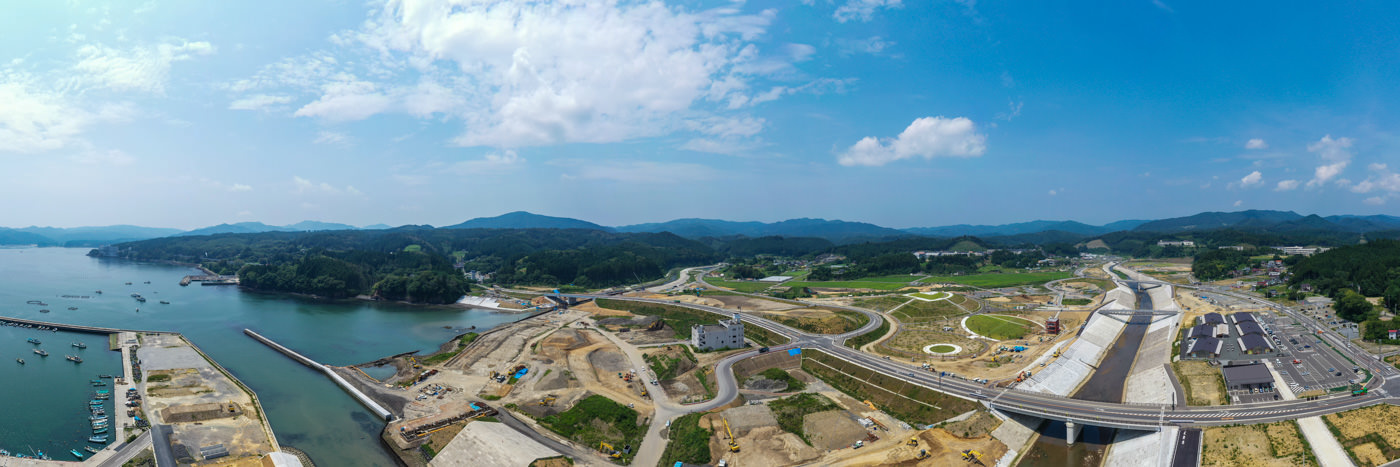 左側に複数の船が停泊している海が広がり、陸地には工事現場や道路、橋などの人工的な構造物が点在し、背景には緑豊かな丘や山々が部分的に雲に覆われた空の下に広がっている、自然と開発が混在する沿岸地域のパノラマ風景の写真