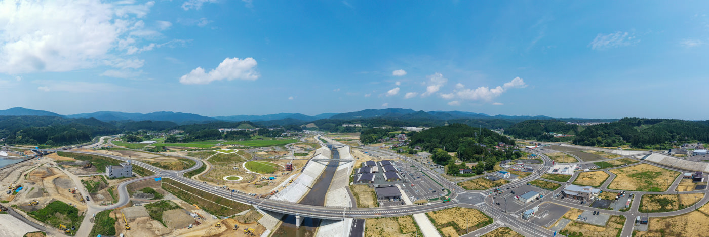 新しく整備された高速道路や道路網が複数の橋や交差点を含んで広がる風景で、周囲には開発中の土地や建物、駐車場が点在し、背景には緑の丘や山々が広がる晴天の写真