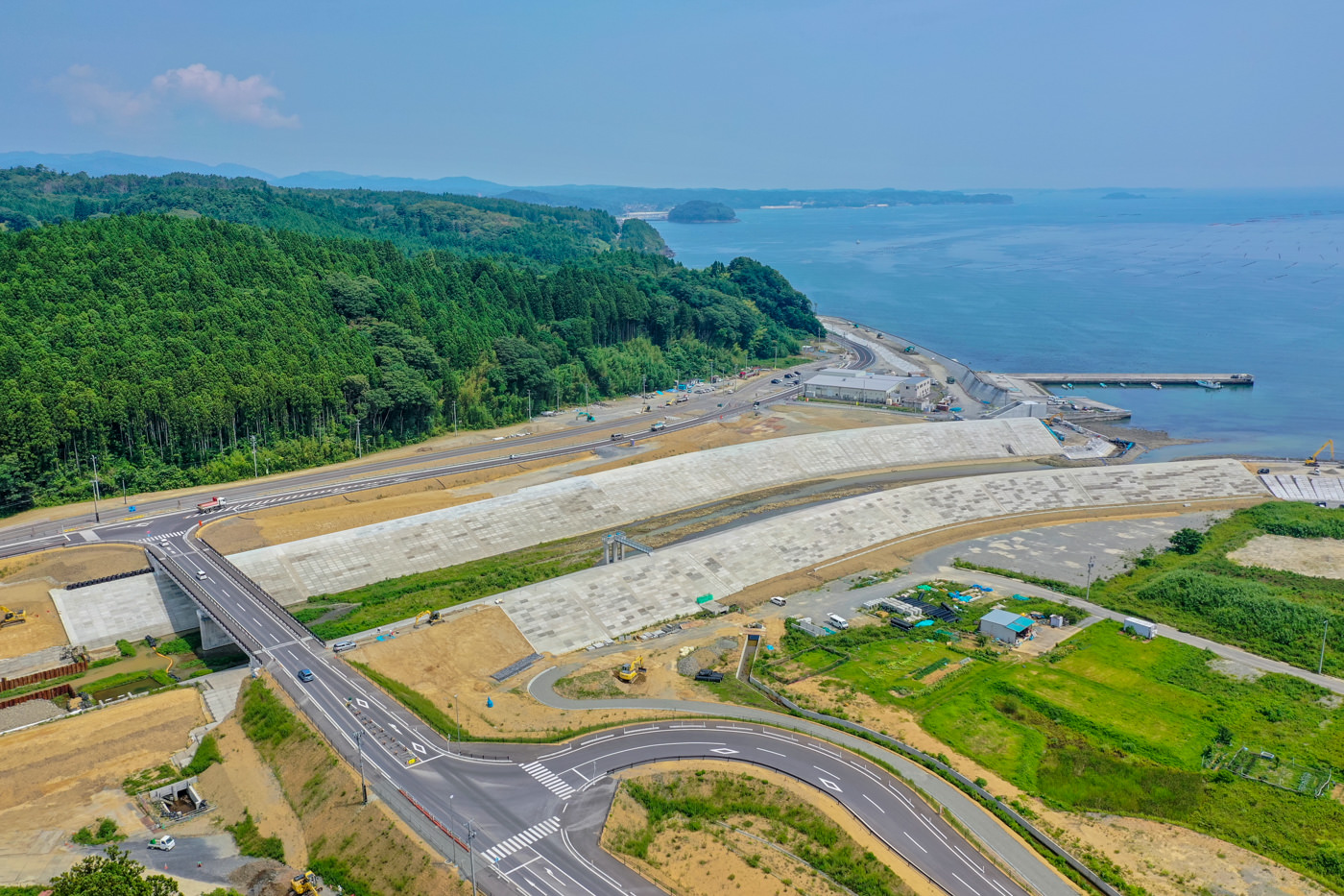 左側に広がる緑豊かな森林と右側に見える海に挟まれた地域に、複数車線の新しく整備された道路が曲線を描いて走り、沿岸には津波や洪水対策と思われるコンクリートの防護施設が設置されている晴天の風景の写真