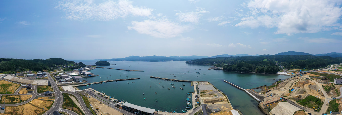 港に複数の船が停泊し、海岸沿いには建物やインフラが整備され、道路や工事現場が広がり、背景には緑の丘が見える部分的に曇った空の下の海辺の風景の写真