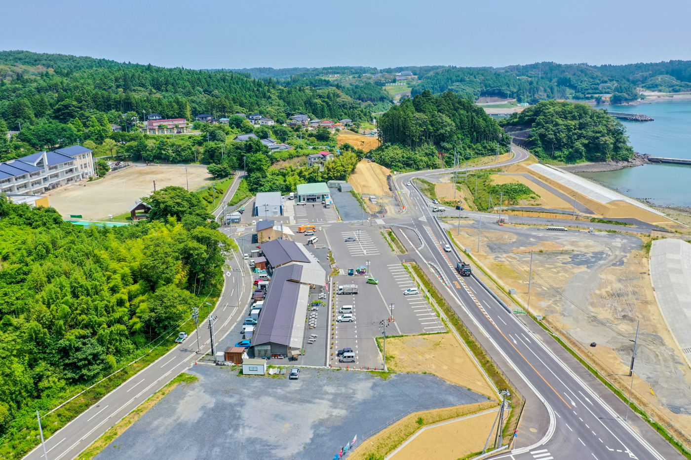 緑に覆われた山々と青く広がる海に囲まれた地域に、複数の車が停まっている駐車場と白い建物が並んでいる様子が見える風景の写真