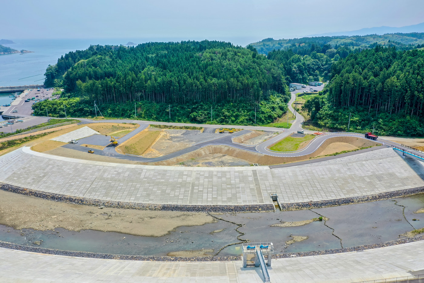 海岸沿いに設けられた高さのあるコンクリート製の防波堤が画面中央に大きく写っており、その背後には緑が濃く生い茂る広大な森林が広がっている風景の写真