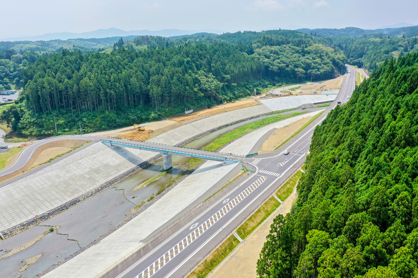 緑に覆われた山々に囲まれた谷間を縫うように高速道路が通っており、その上を橋が横切っている静かな自然の中の交通インフラの風景の写真