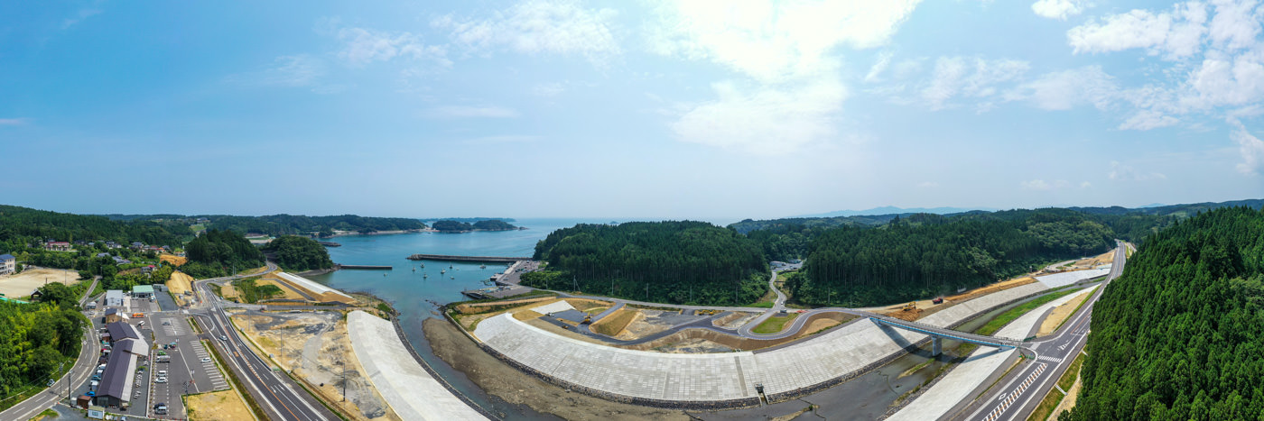 青く澄んだ空の下、緑に覆われた山々に囲まれた海岸線が広がり、その手前にはコンクリートで造られた防波堤と整備された道路が続いている風景の写真