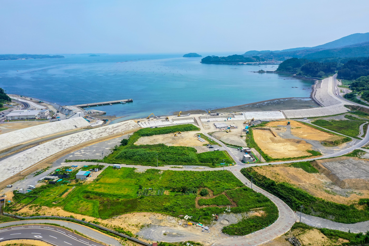 海岸沿いに広がる緑の草地と工事現場、道路が入り混じった地域を上空から見下ろした風景で、背景には海と小さな島々が見え、沿岸には防波堤と思われるコンクリート構造物が並び、建物や車両が点在し、遠くには丘や山々が広がっている写真