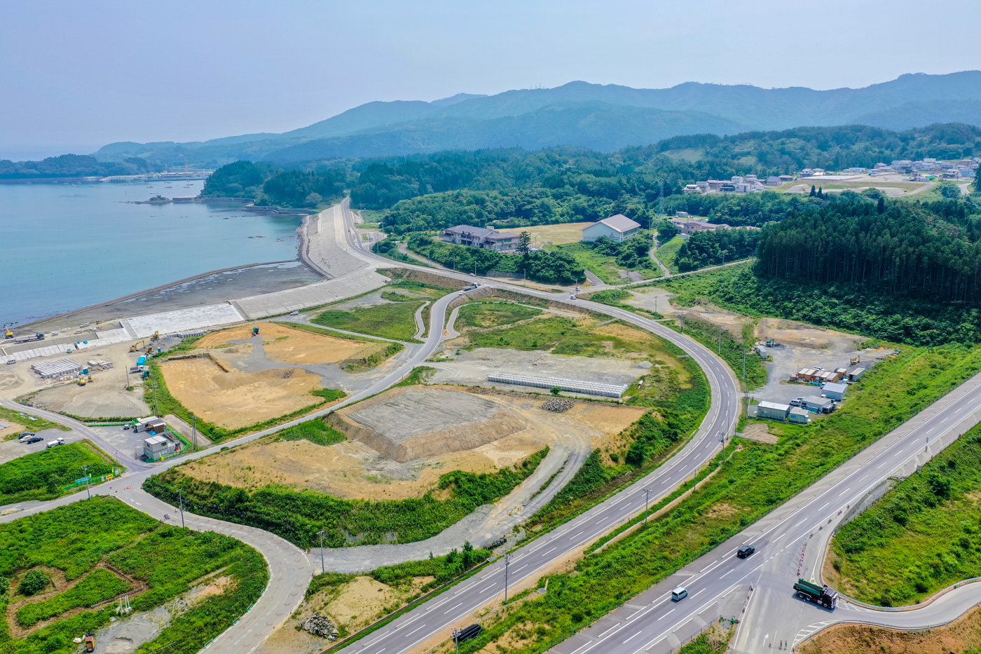 左側に海岸線が広がり、緑の草地と建物が点在する地域を上空から見下ろした風景で、曲がりくねった道路が土地を横断し、遠くには山々がそびえ、いくつかの区画では開発や工事が進められている様子が見える写真