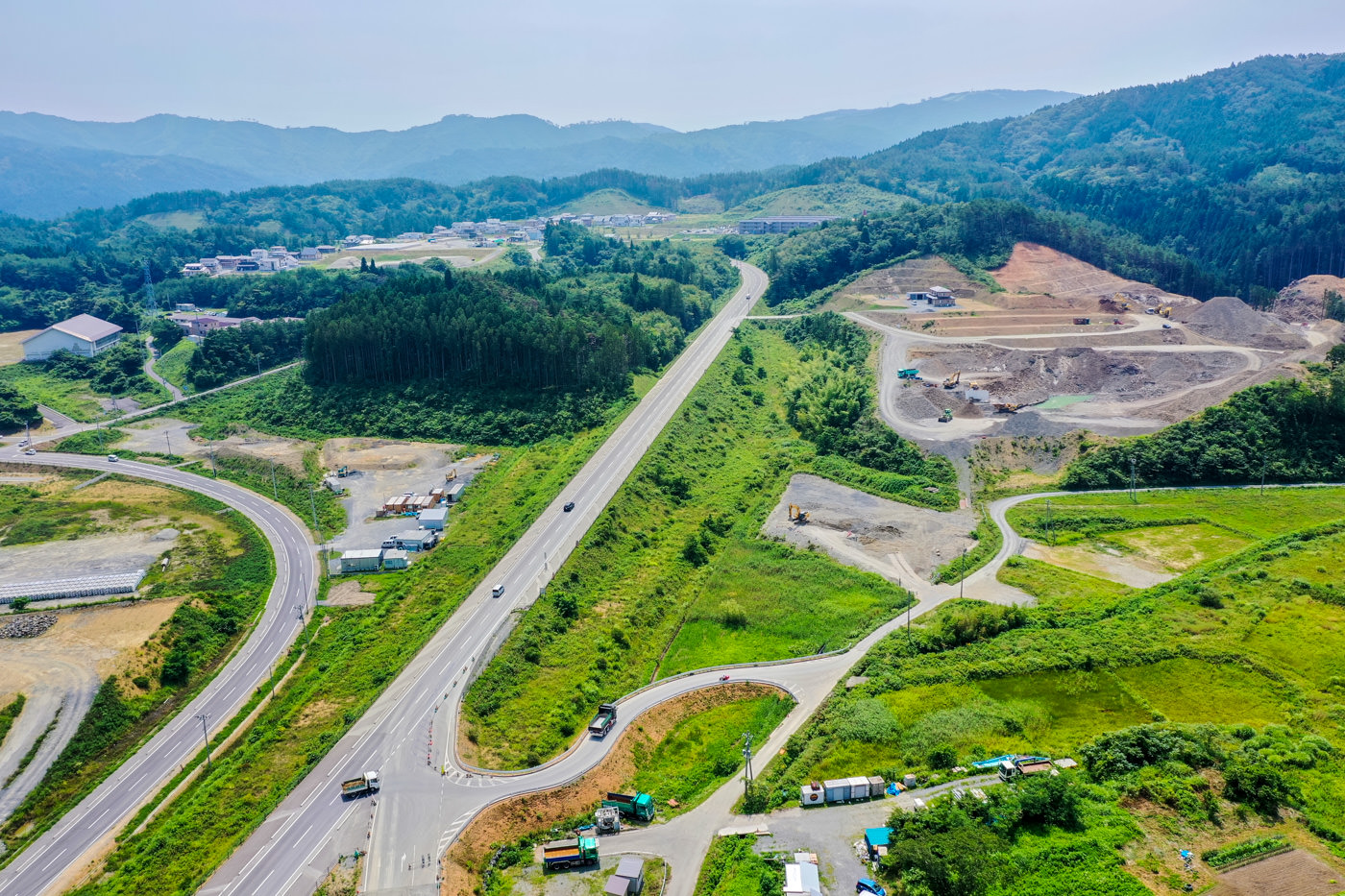 緑の草地と丘陵が広がる農村地域を上空から見下ろした風景で、複数の道路が交差し、車両が走行しており、建物が点在し、背景には森林に覆われた山々が広がる晴天の写真