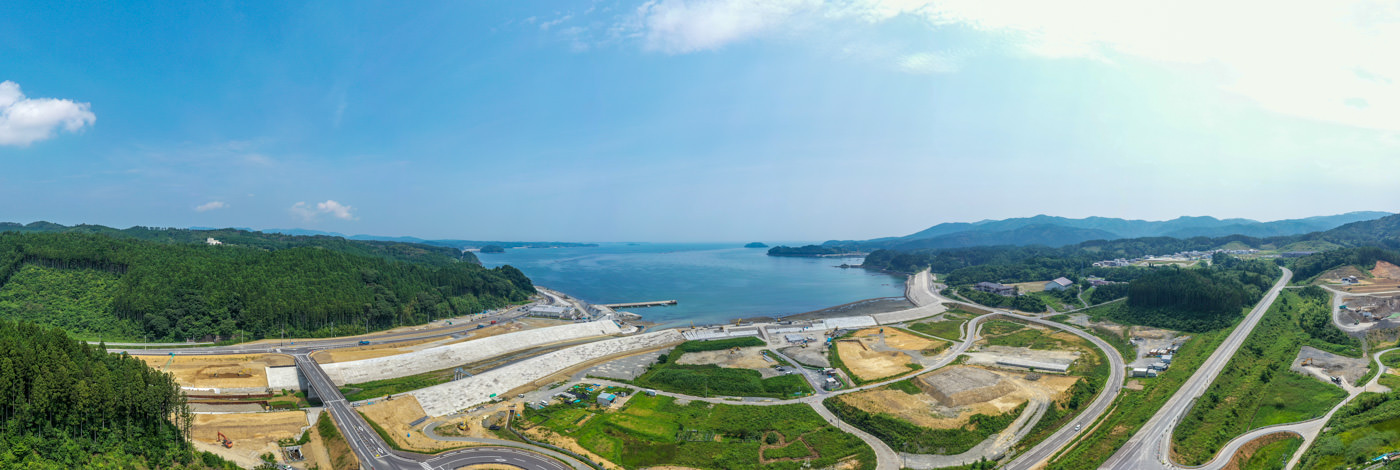海岸沿いに広がる緑の森林と道路、複数の工事現場が見える地域を広く見渡す風景で、穏やかな海と雲のある青空が背景に広がっている写真