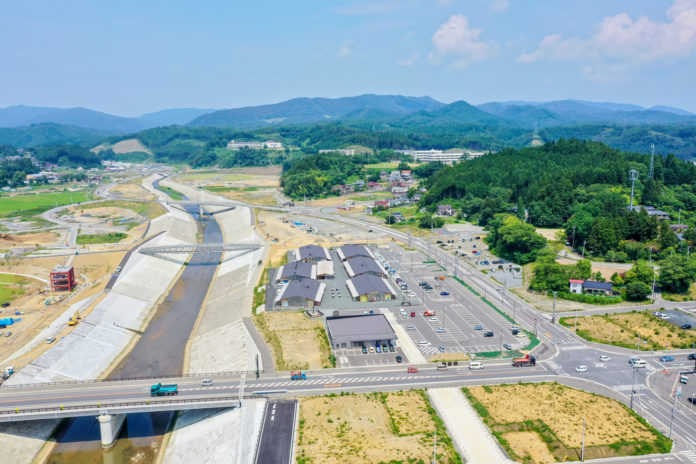 コンクリートで護岸された川に橋がかかり、その周囲に建物や駐車場、車が走る道路が整備され、背景には緑豊かな山々が広がる風景を上空から見下ろした写真