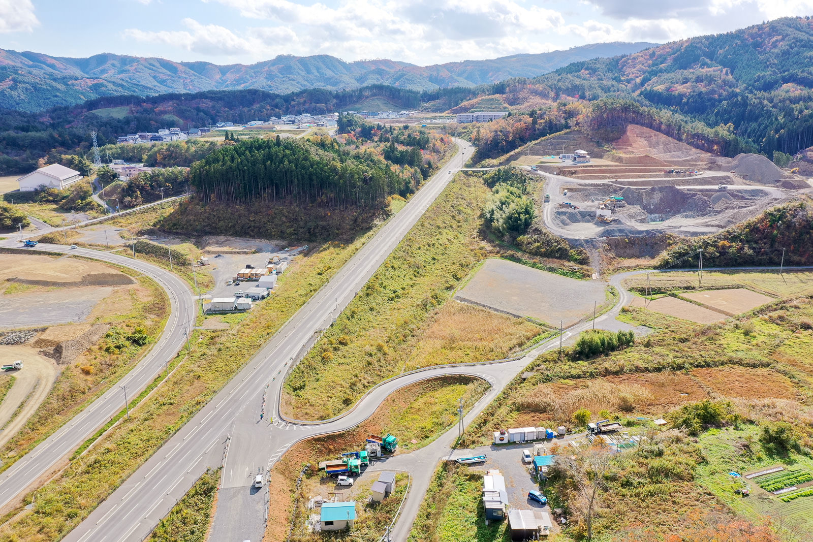 丘陵地帯に広がる田園風景を空から捉えた構図で、幹線道路と枝道が網の目のように走り、道路沿いには車両が停まり、建物が点在し、森林と開けた土地が混在する中、背景には山々が広がる地方の風景の写真