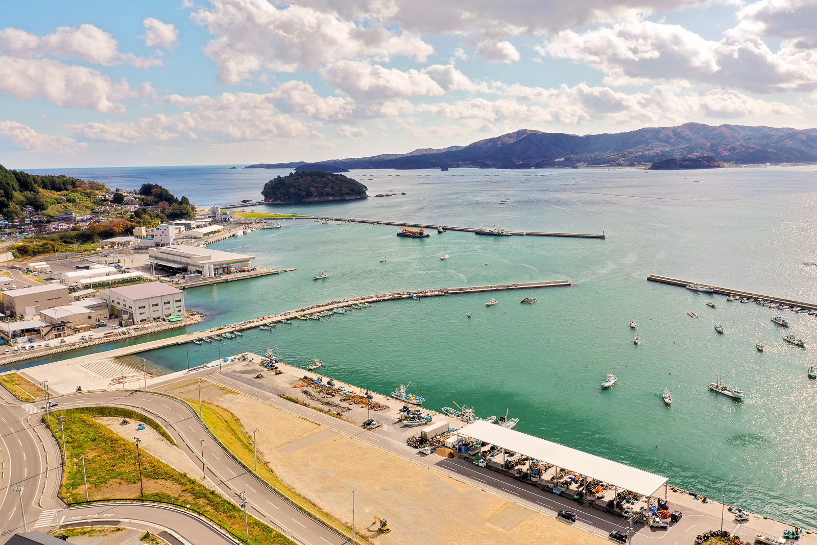 青緑色の海に複数の桟橋が伸び、船が停泊する港が広がり、周囲には建物や道路、駐車場が点在し、左側には緑地があり、背景には丘や山々が見え、空には雲が浮かぶ海岸地域の空撮の写真