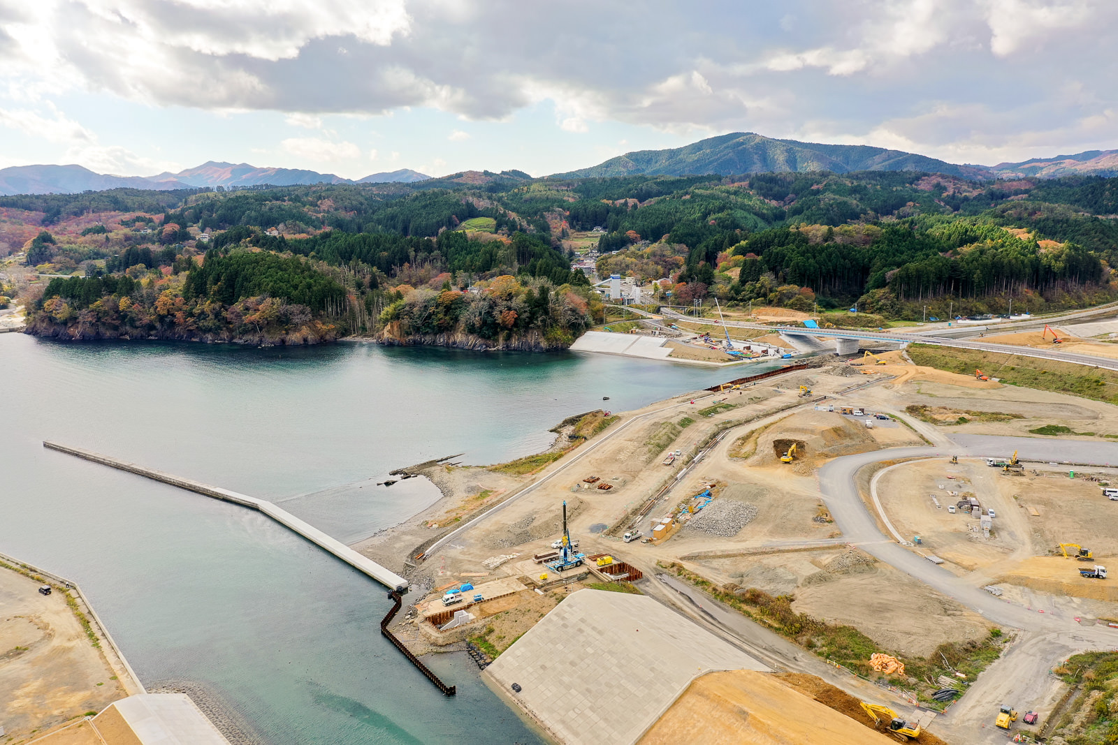 海に長い桟橋が突き出した沿岸の工事現場を空から見下ろした風景で、周囲には工事用の機材や車両、資材が並び、背景には木々に覆われたなだらかな丘が広がる空撮の写真