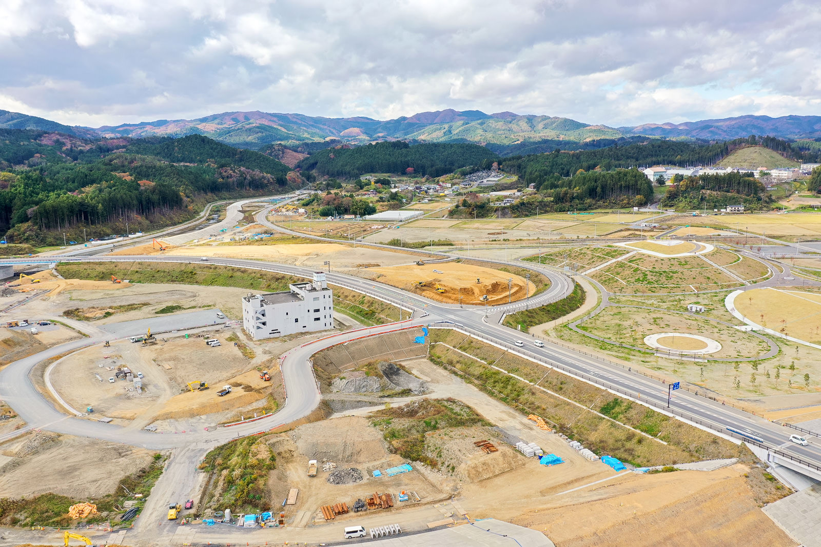 中央に白い建物があり、その周囲に道路や通路が整備されつつある広大な工事現場が広がり、土や建設資材が点在し、背景にはなだらかな丘と山々が広がり、空には雲が浮かぶ工事現場の空撮の写真