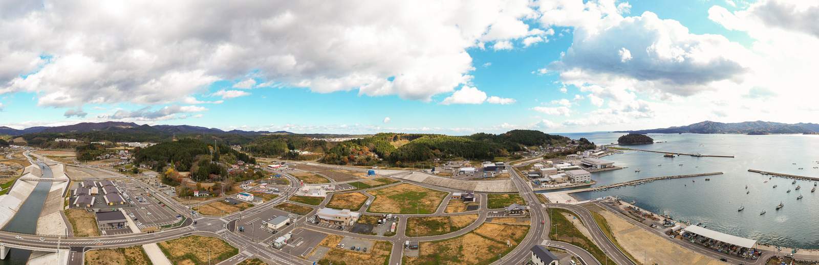 海沿いの町を空から見下ろした風景で、手前には道路や建物、駐車場が広がり、奥には緑豊かな丘や森林が続き、海岸には船が停泊する港があり、空には雲と青空が見える空撮の写真