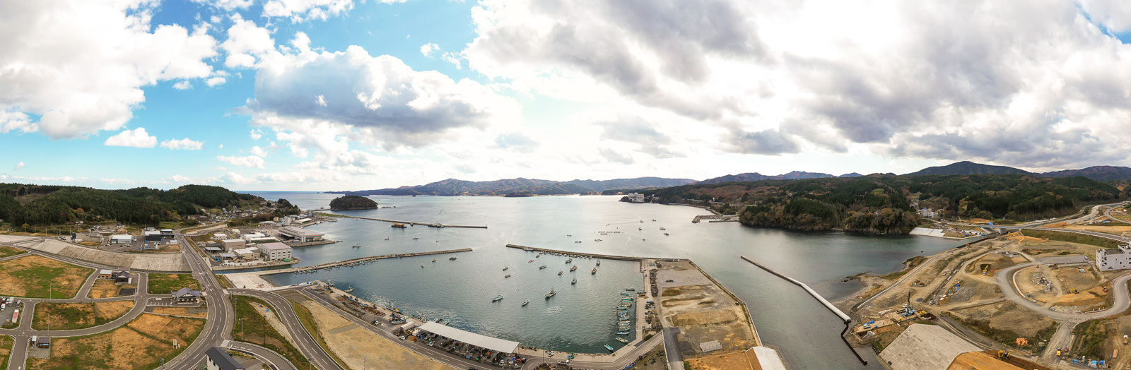 港に複数の桟橋が海へ伸び、たくさんの船が停泊している海岸地域を広く見渡した風景で、周囲には建物や道路、工事現場が点在し、背景には丘や山々が広がり、空には雲が浮かぶ海辺の空撮の写真