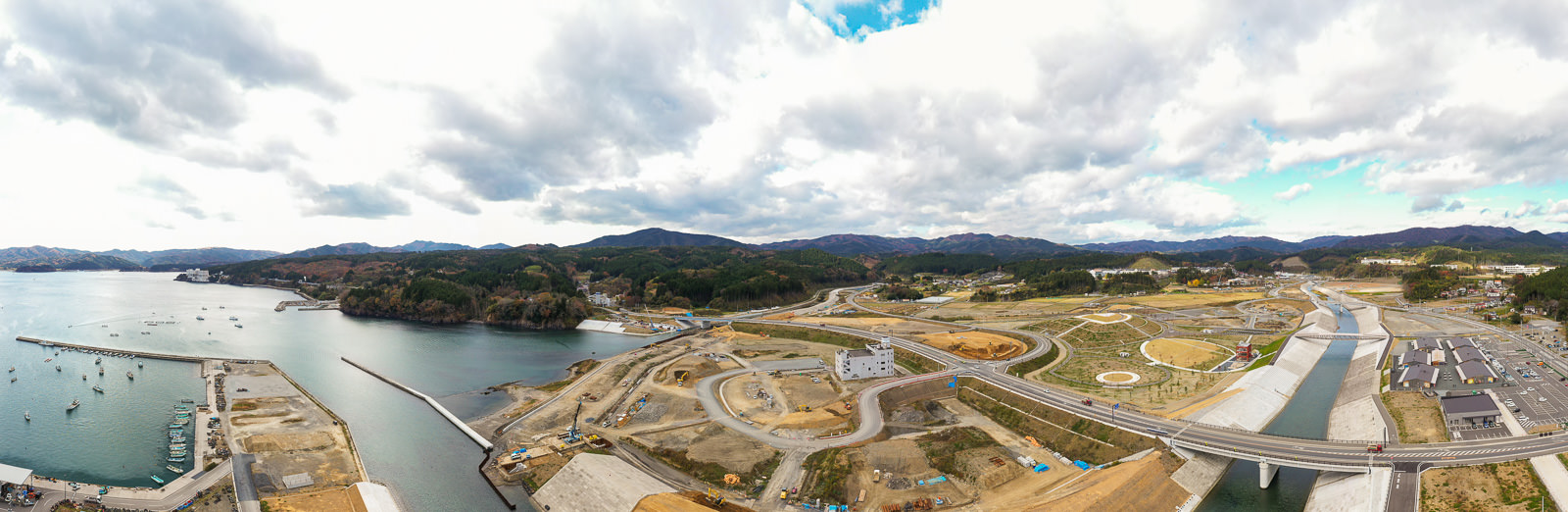 左側に複数の船が停泊する港があり、周囲には工事現場や道路、橋が広がり、背景には丘や山々が連なり、空には雲が浮かぶ海岸地域の空撮の写真